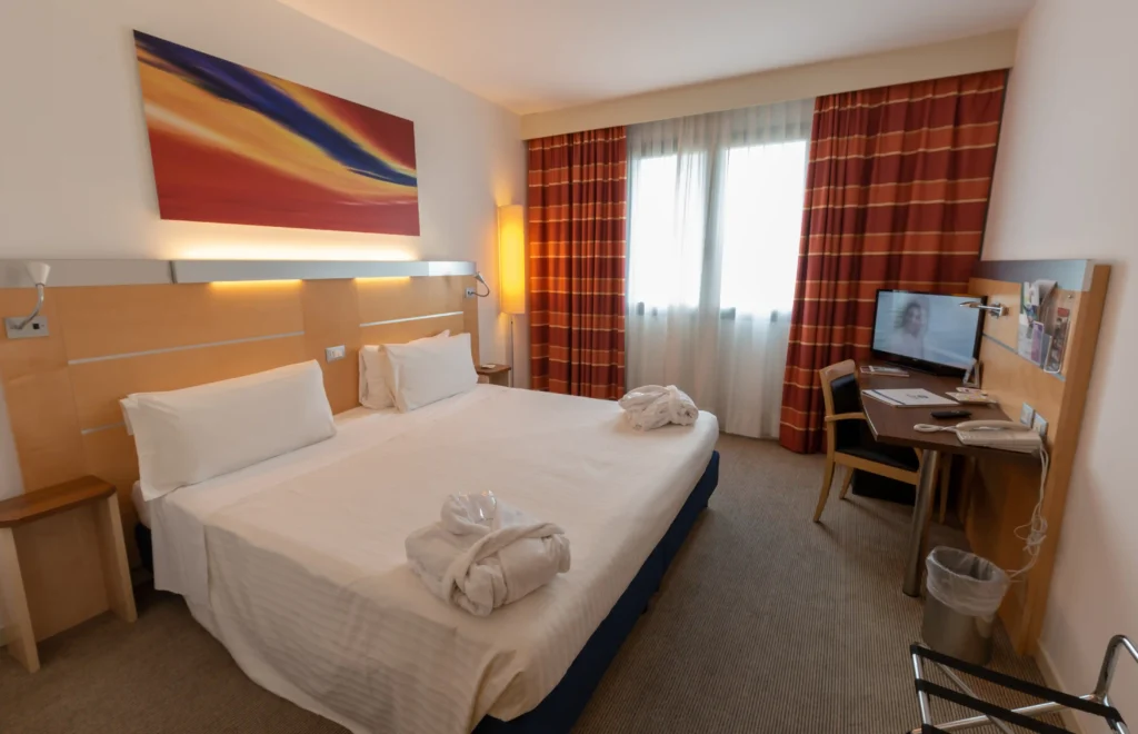 In the picture, the Superior Room of the Palace Inn Hotel: we can see the double King bed with two bathrobes on it. On the right, a desk with a TV. In the background, a window with closed curtains.