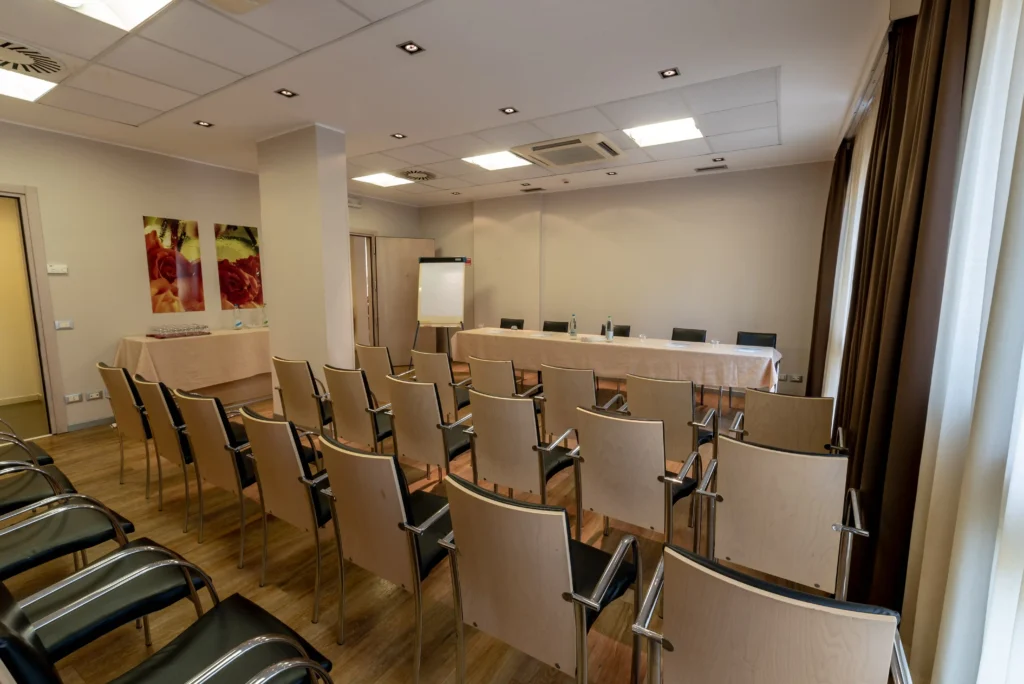 Nell'immagine, la sala meeting Eridano del Palace Inn Hotel: si vedono alcune file di sedie e, di fronte, il tavolo dei relatori con di fianco una lavagna a fogli mobili.