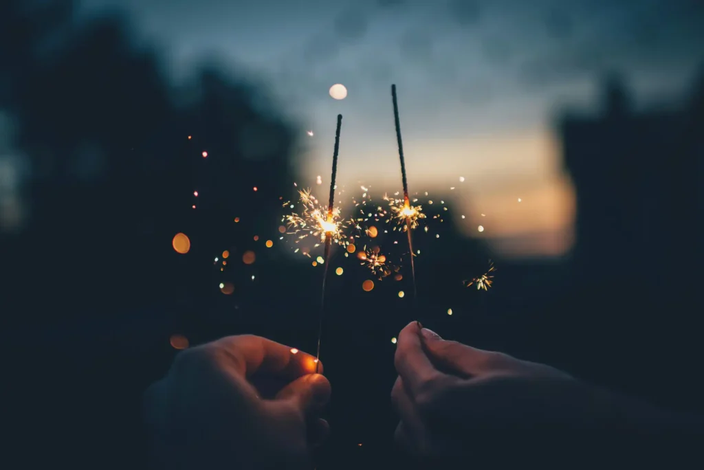 In the picture, a close-up on the hands of two people holding two sparkler fireworks.