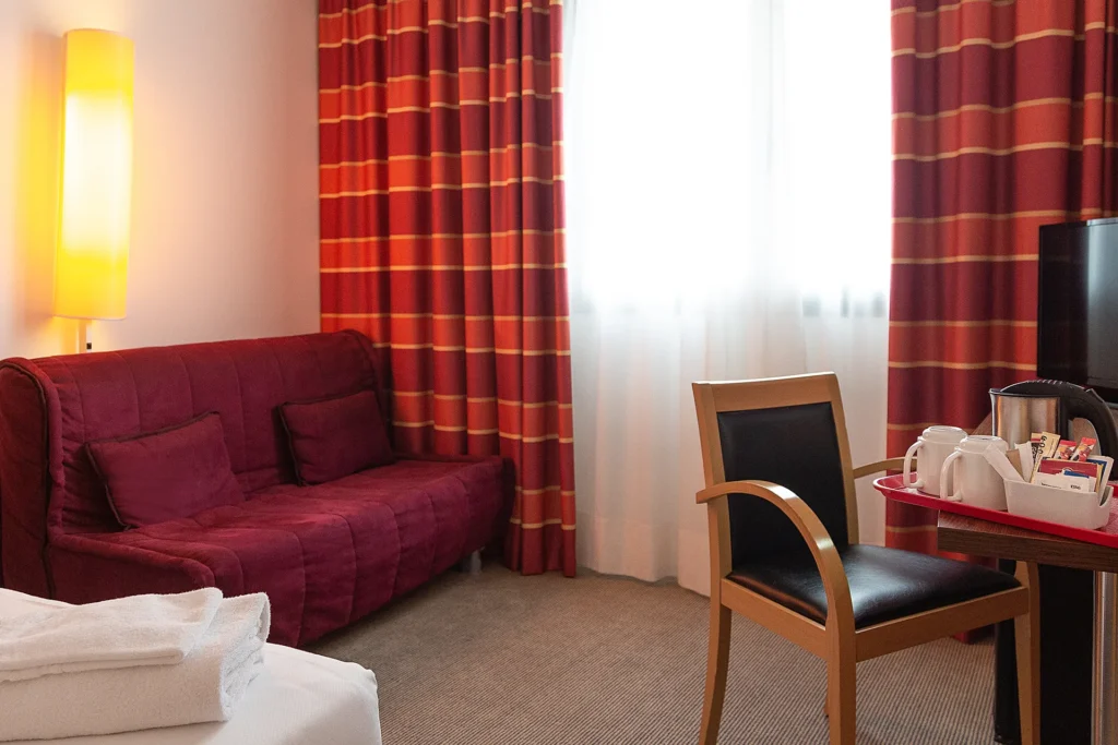 Nell'immagine, la camera Standard del Palace Inn Hotel: si vedono un divano sulla sinistra e un tavolo con una sedia sulla destra: sul tavolo, è poggiato un vassoio con un bollitore e due tazze. Si intravede anche una TV.